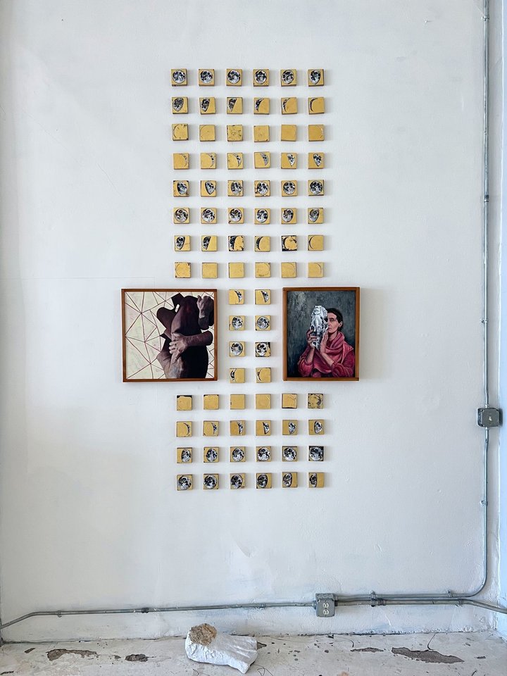 Installation view: dozens of small gold-ground square panels painted with moon phases, arranged in a tall grid on a white wall around two framed paintings — a figure in a wide hat, and a portrait of a woman in a red shawl holding a carved head.