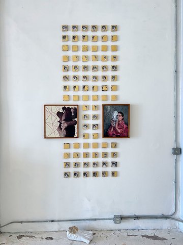 Installation view: dozens of small gold-ground square panels painted with moon phases, arranged in a tall grid on a white wall around two framed paintings — a figure in a wide hat, and a portrait of a woman in a red shawl holding a carved head.