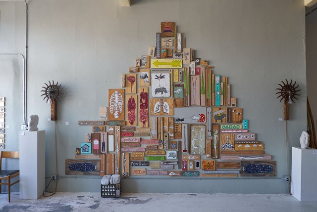 Installation view: dozens of small paintings on found wood assembled into a triangular, pyramid-shaped arrangement on a sage-grey wall, flanked by two spiked metal sconces, with small carved sculptures on white plinths at either side.