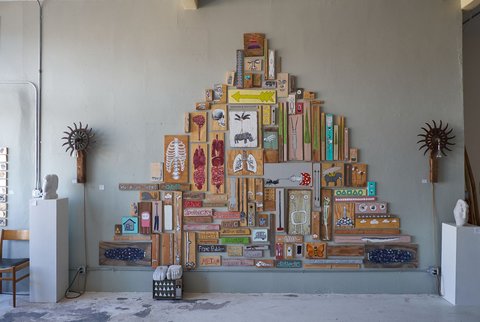 Installation view: dozens of small paintings on found wood assembled into a triangular, pyramid-shaped arrangement on a sage-grey wall, flanked by two spiked metal sconces, with small carved sculptures on white plinths at either side.