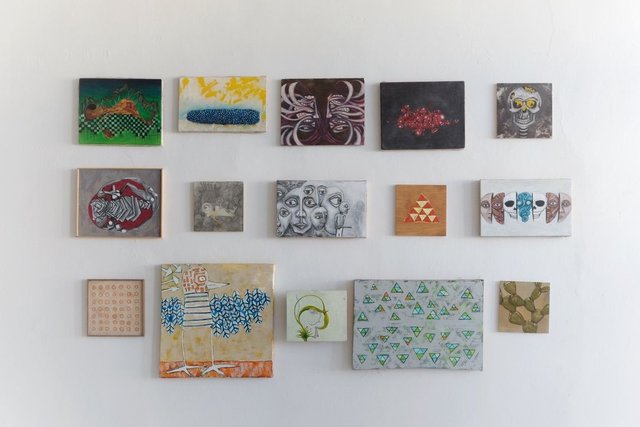 Installation view of fifteen small paintings hung in three loose rows on a white wall — among them a checkered landscape, a blue mass on yellow, a crowd of faces, an orange triangle of triangles, and a panel of green triangles.