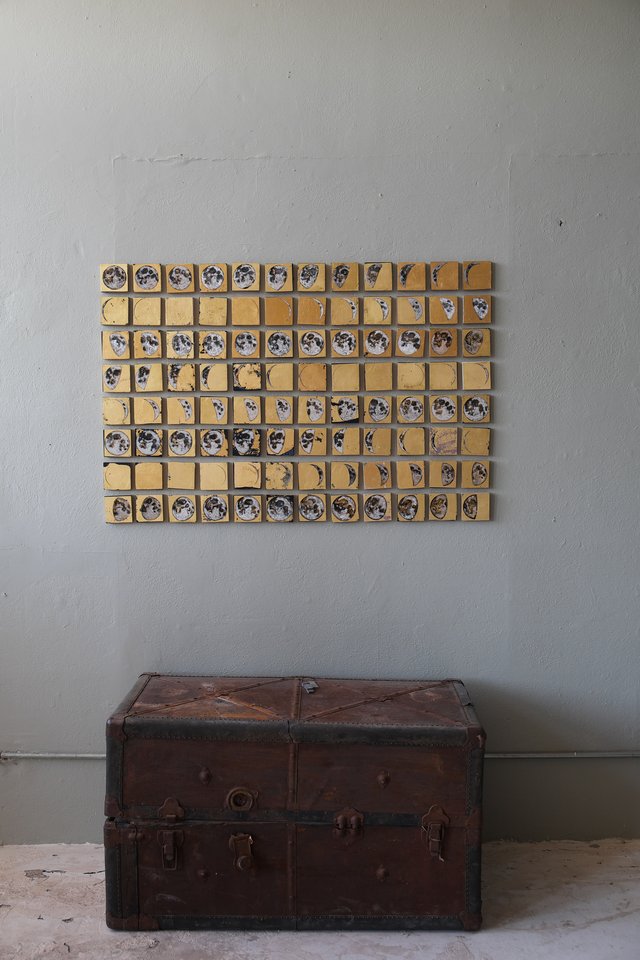 Grid of small square paintings on 24kt gold leaf depicting a summer's worth of moon phases, mounted on a gray wall above a vintage leather trunk