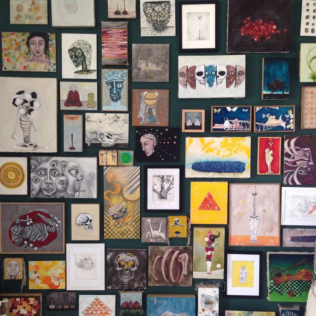 Salon-style hang on a deep green wall, densely covered with dozens of small paintings and framed drawings edge to edge — faces, skulls, a bird skull, geometric heads, botanical studies and abstract panels in mixed sizes and frames.