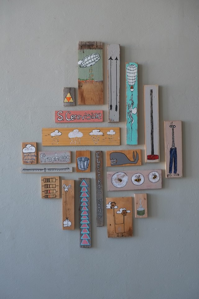 Installation view: a cluster of small paintings on scraps of found wood arranged on a pale grey wall — an airship, a hot-air balloon, a whale, three bees, a rain cloud, a row of triangles — interspersed with hand-lettered boards reading “Prophets”, “S. Cerevisiae” and “Electric Wind”.