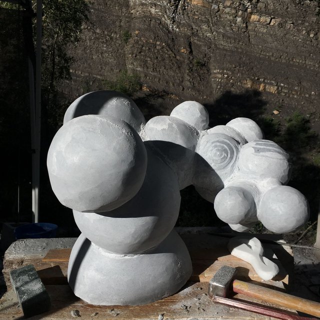 Carved white marble sculpture in progress on an outdoor workbench: a cluster of joined spheres and rounded masses, one face incised with concentric rings, with mallets and a small carved offcut beside it and a stratified rock cliff behind.
