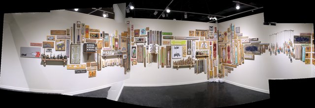 Panoramic installation view of a gallery: a long white wall densely hung with more than a hundred small paintings on found wood, shelved objects and framed pieces, wrapping around a corner to a row of hanging strings on the right.