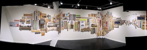 Panoramic installation view of a gallery: a long white wall densely hung with more than a hundred small paintings on found wood, shelved objects and framed pieces, wrapping around a corner to a row of hanging strings on the right.