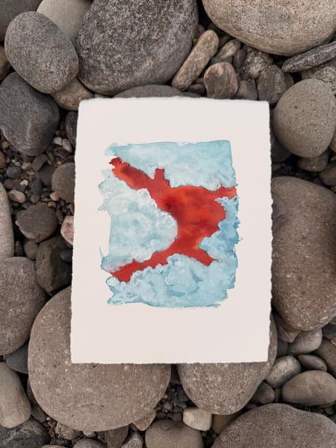 Watercolour on a deckle-edged sheet laid on smooth river cobbles: pale blue cloud shapes around a branching, vivid red-orange form that crosses the sheet like a gap in the sky.