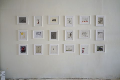 Installation view of eighteen small white-framed prints hung in three even rows of six on a white plaster wall, the images ranging from botanical studies to figures and abstract marks.